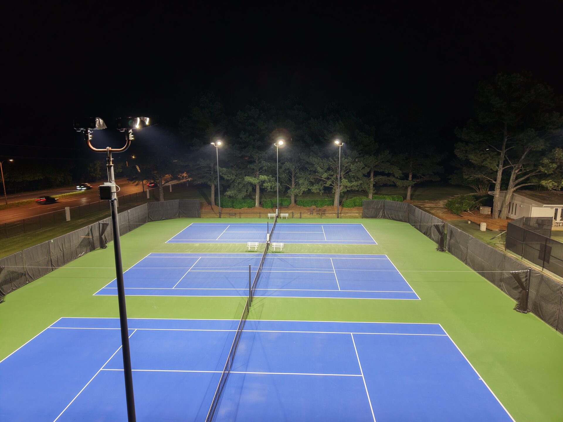 Ground-level player perspective view down the length of the tennis courts, showing even illumination with no dark baseline areas
